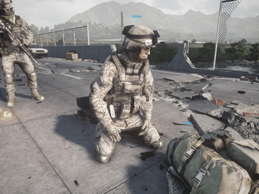 A soldier in modern combat gear kneels on a destroyed bridge, tending to a fallen comrade in Battlefield 3. The background features mountainous terrain and broken infrastructure, emphasizing the realism of the warzone. Purchase Battlefield 3 Limited Edition PC EA Play Origin CD Key at RushGame.co