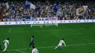 In-game moment from EA Sports FC 24 Ultimate Edition on Xbox One & Xbox Series X/S, featuring a powerful shot from a Real Madrid player in a UEFA Champions League match. The goalkeeper dives in an attempt to stop the ball, while the crowd waves white and blue flags in support. Get your EA Sports FC 24 Ultimate Edition CD Key at RushGame.co for instant play